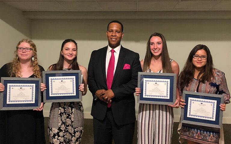 Five individuals stand indoors, four holding framed certificates. The person in the center wears a suit, reminiscent of a formal Florida gathering, while the others sport semi-formal attire. A plain wall serves as the understated backdrop to this distinguished moment.