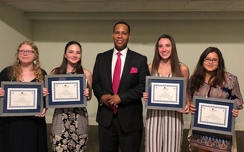 Five individuals stand indoors, four holding framed certificates. The person in the center wears a suit, reminiscent of a formal Florida gathering, while the others sport semi-formal attire. A plain wall serves as the understated backdrop to this distinguished moment.
