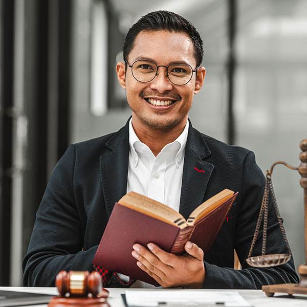 A man wearing glasses and a suit sits at his desk with a legal book, gavel, and scales of justice, smiling warmly at the camera. His professional demeanor reflects the expertise common among Florida's esteemed legal professionals.