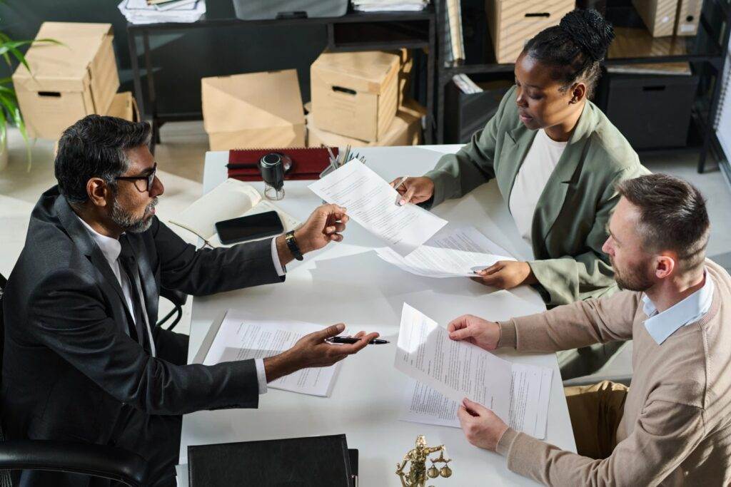 Three people sit around a table in a bustling Florida office, engaged in lively discussion over documents. Surrounded by boxes and office supplies, the setting buzzes with the energy of productivity and warm sunshine seeping through the windows.