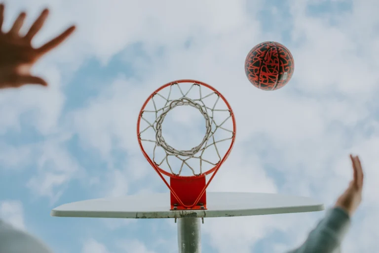 A basketball is mid-air approaching a hoop, viewed from below against a cloudy Florida sky. Two hands reach towards the ball.