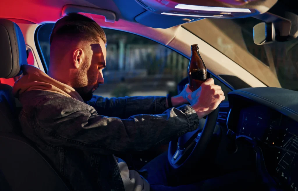 A man in a car holds a beer bottle with both hands on the steering wheel. The interior, reminiscent of a neon-drenched Florida night, is dimly lit with pink and blue lights.