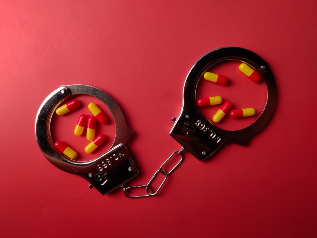 A pair of metal handcuffs with red and yellow capsules inside each cuff, set against a solid red background, evokes the concept of constructive possession often cited in Florida law cases.