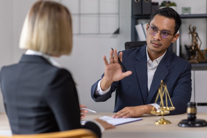 A man in a suit gestures while speaking to a woman across a desk with legal documents, a gavel, and scales of justice—discussing when to call a lawyer for misdemeanor charges.