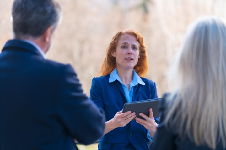 A woman in a blue suit holds a tablet and speaks to two people outdoors, discussing when to call a lawyer after facing a misdemeanor charge.