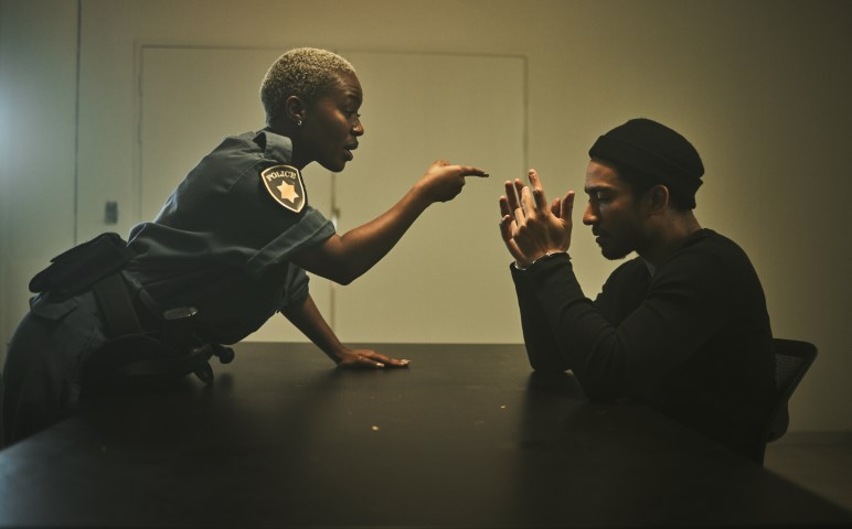 A police officer leans over a table, pointing and speaking to a man in a black shirt and beanie, who sits with his hands clasped and head lowered, seemingly struggling to respond to a false accusation of crime.
