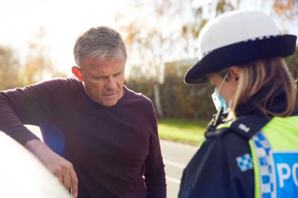 A man in a maroon sweater stands next to a police officer outdoors on a sunny day, engaged in conversation and possibly lying to the police.