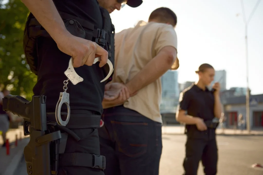 Police officers detain a person outdoors; one officer holds handcuffs while another radios in the background after the suspect is accused of lying to the police.