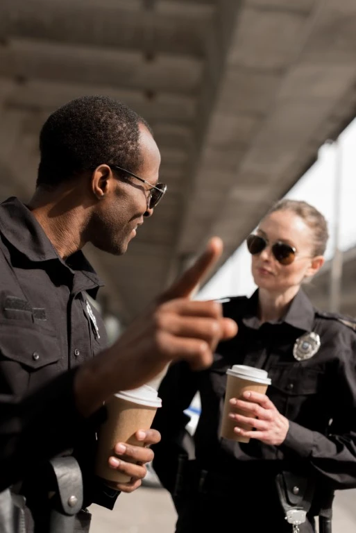 Two police officers in uniform, each holding a coffee cup, are standing outdoors under a bridge, discussing the challenges of spotting when someone is lying to the police.