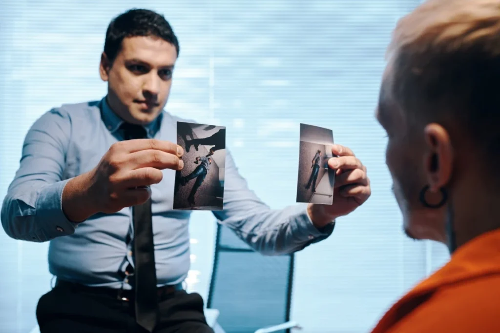 A man in a blue shirt shows two photographs to another person during an interview or interrogation, possibly investigating whether someone is lying to the police in an office setting.