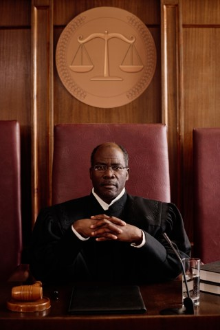 A judge sits at a courtroom bench with hands clasped, wearing a black robe. A gavel, documents, and a glass of water are on the desk—reminding you it's wise to know when to call a lawyer if facing a misdemeanor charge.