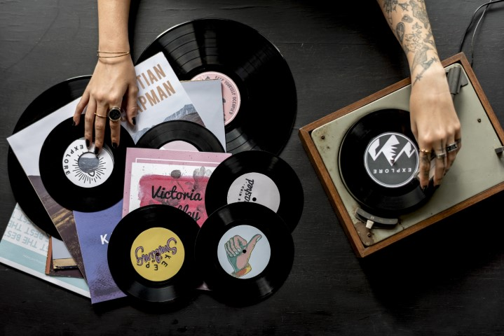 Tattooed arms of artists place a vinyl record on a turntable, with colorful album covers and other records scattered across the table—capturing the creative spirit behind music and entertainment contracts.